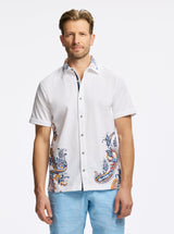 Man wearing a white short-sleeve shirt with colorful patterns and light blue pants on a white background